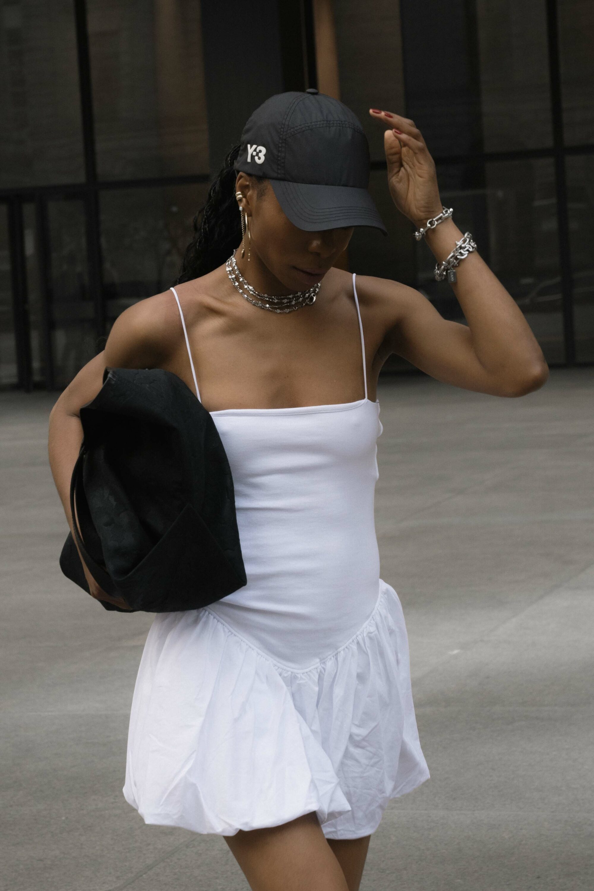 A woman in a white tennis dress, black sneakers, an oversized black tote bag, baseball cap, and silver jewelry smiles as she walks down the street.