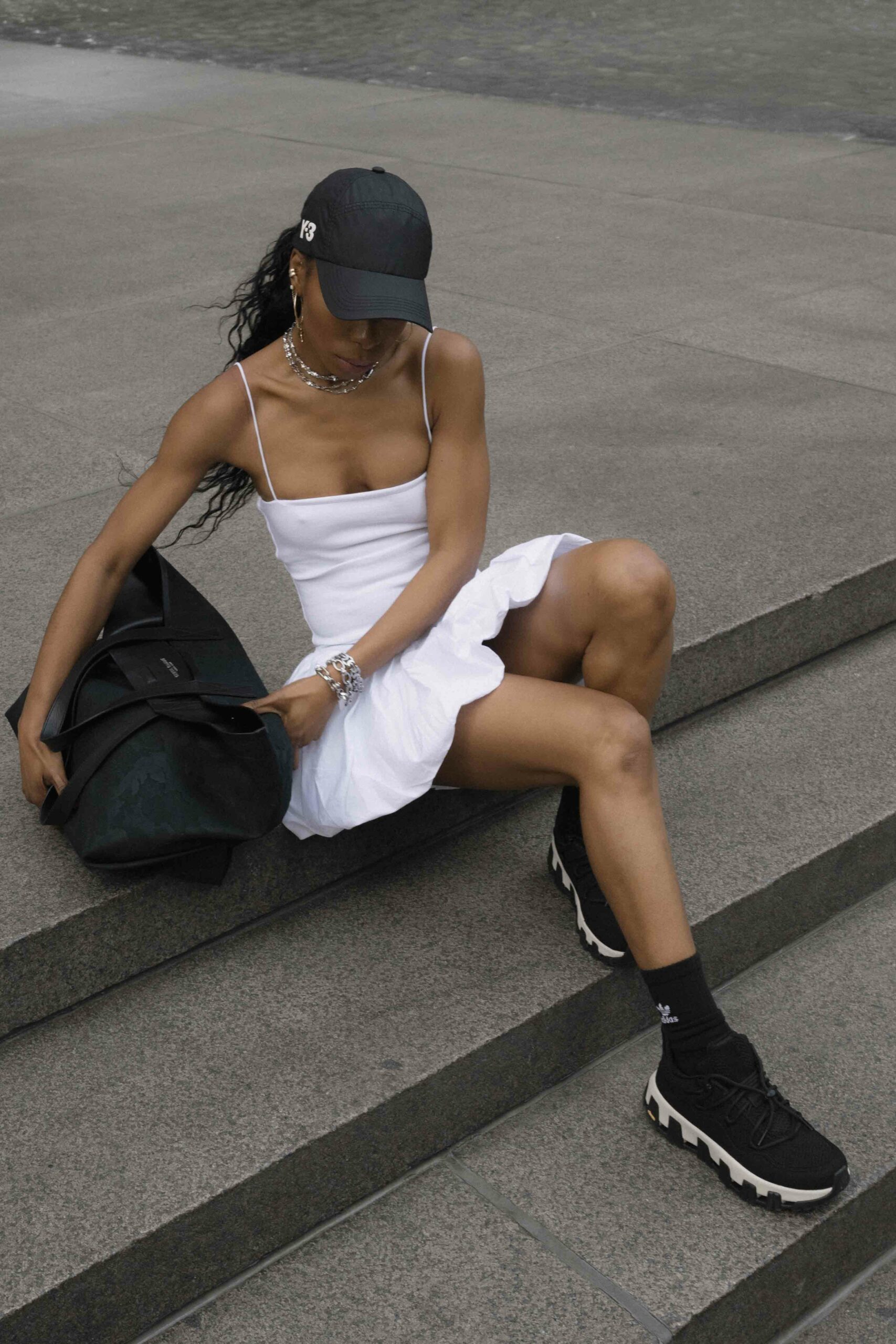 A woman in a white tennis dress, black sneakers, an oversized black tote bag, baseball cap, and silver jewelry sitting on stairs in NYC